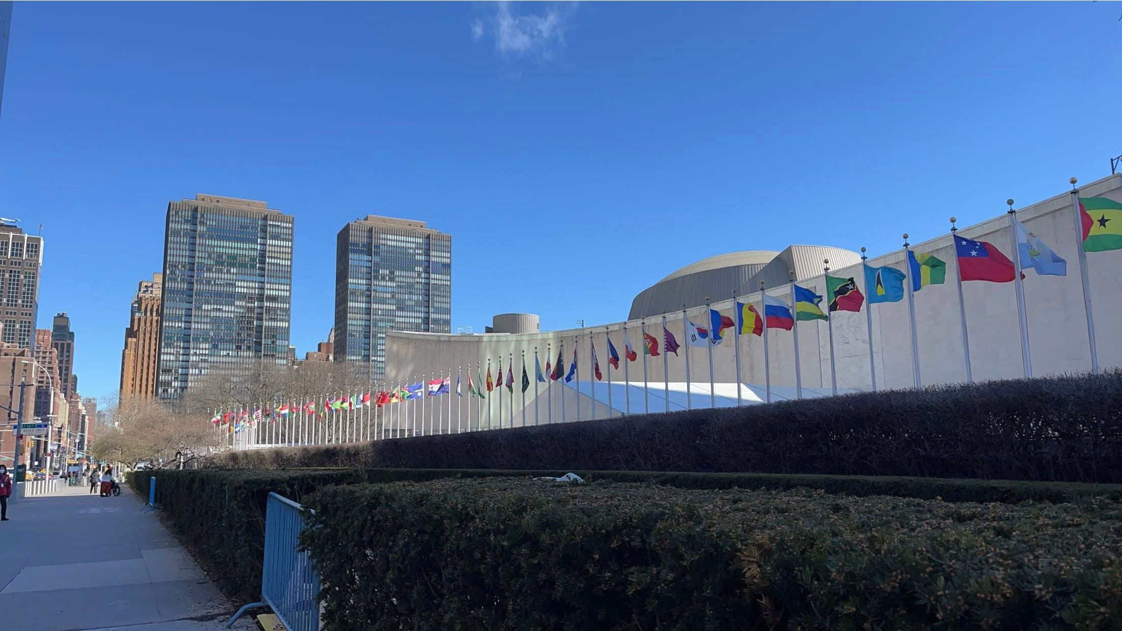Row of international flags flying before the United Nations building and New York City skyscrapers.