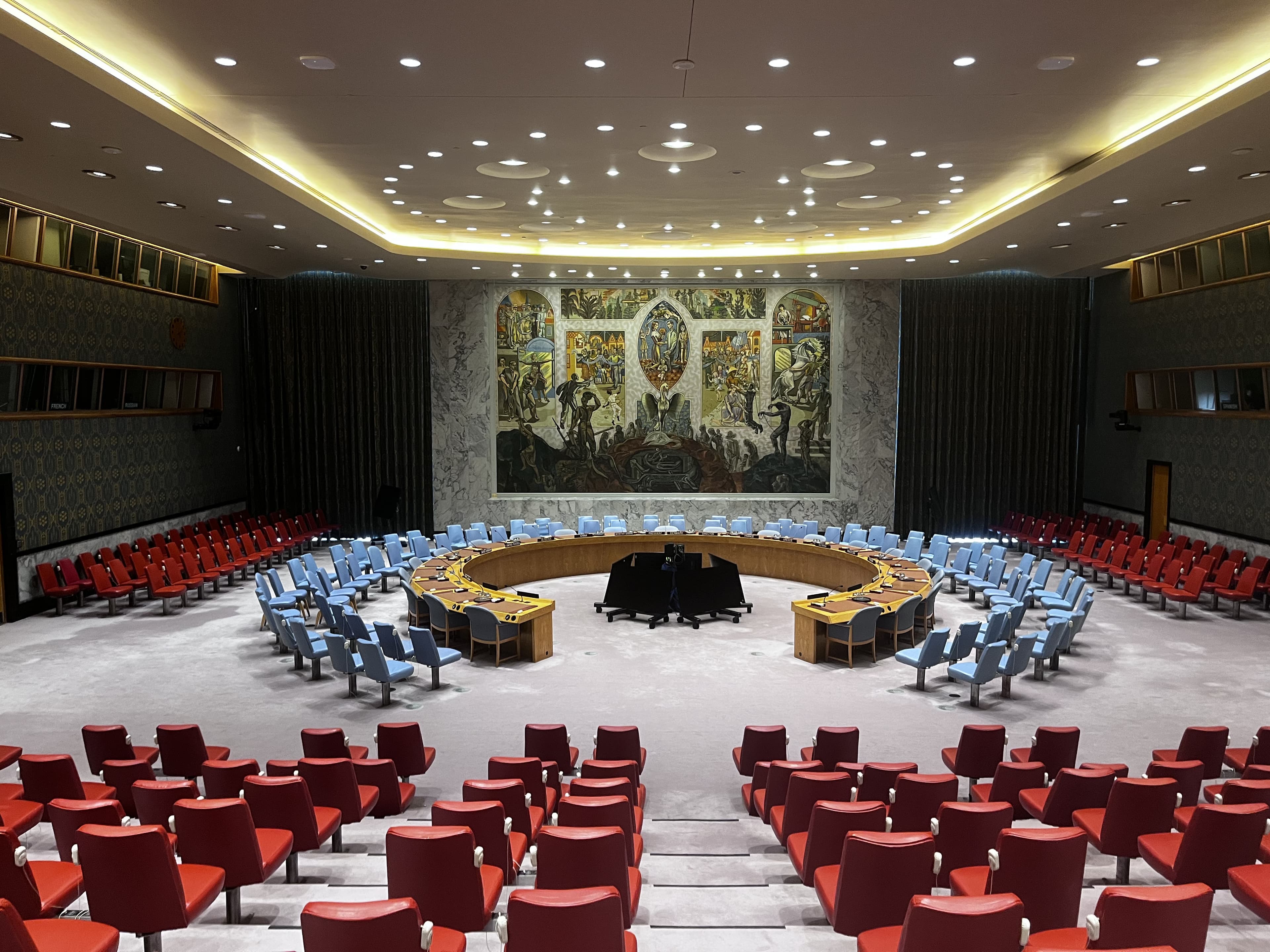 United Nations Security Council chamber with a circular table, blue chairs, and large mural.